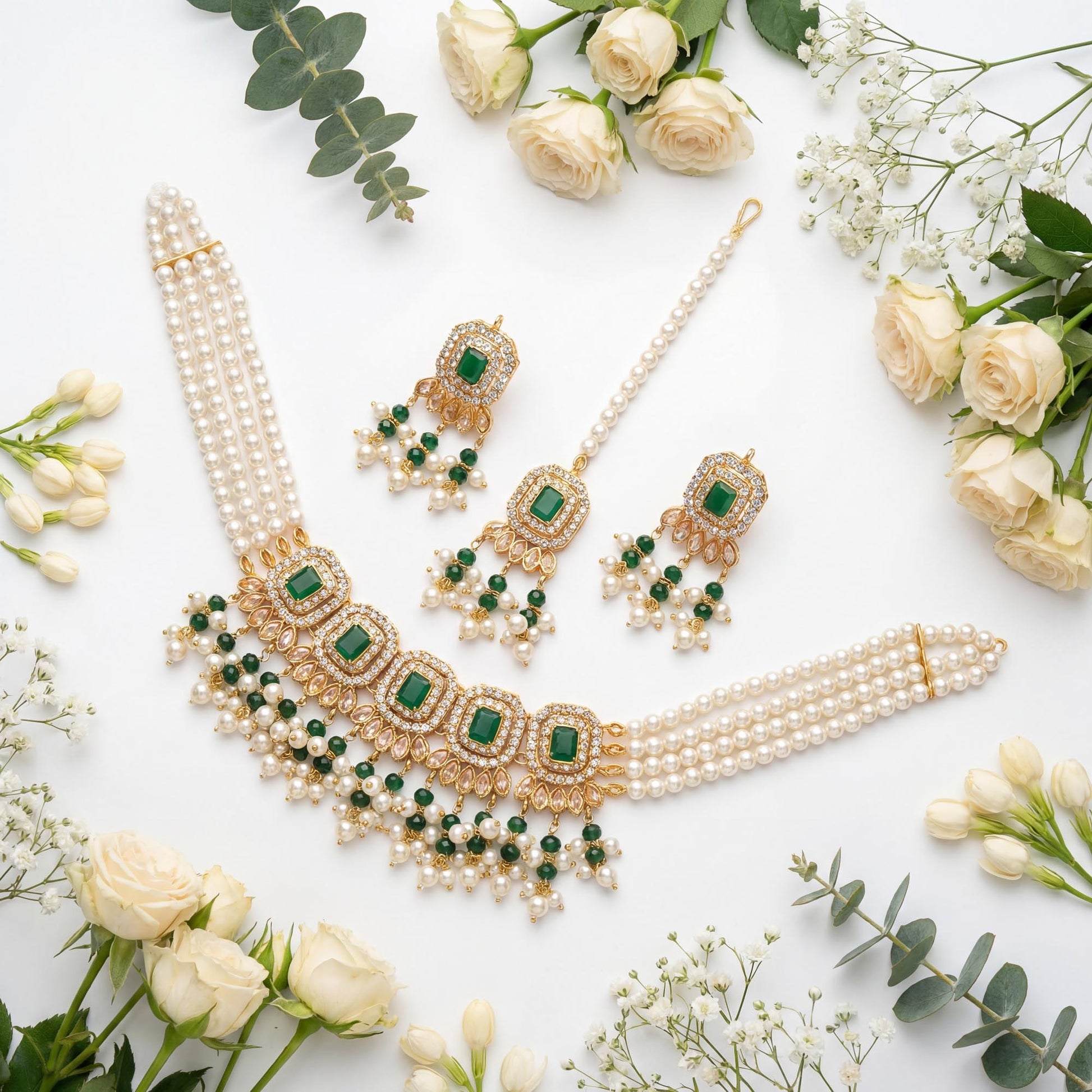 Pearl necklace with green gemstones, matching earrings and a maang tikka on a white background with flowers.