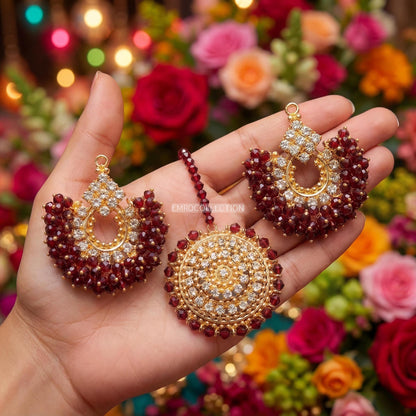 Hand holding maroon chand bali earrings and tikka with a colorful floral background