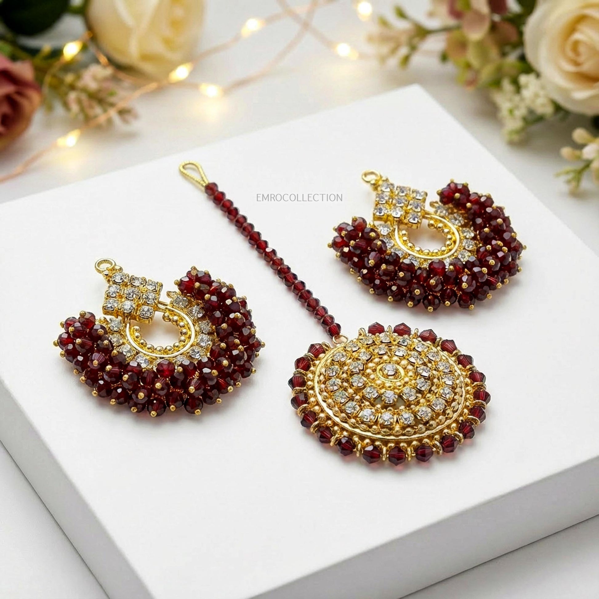 maroon and golden hue chand bai earrings tikka on a white surface with floral decorations.