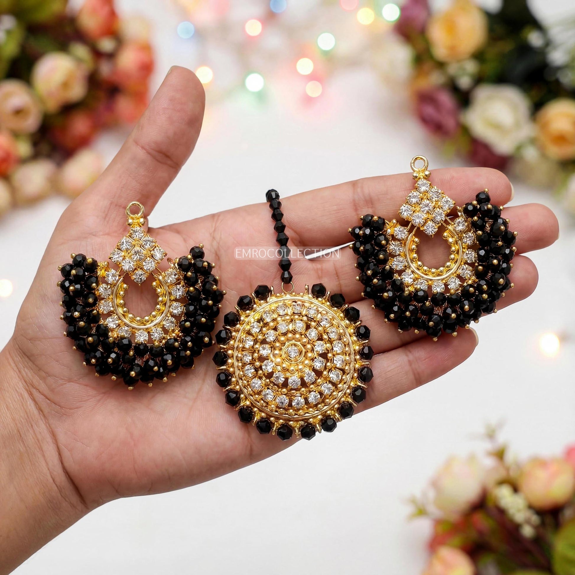 Hand holdin a pair of gold and black chand bali earrings and a matching maang tikka. 