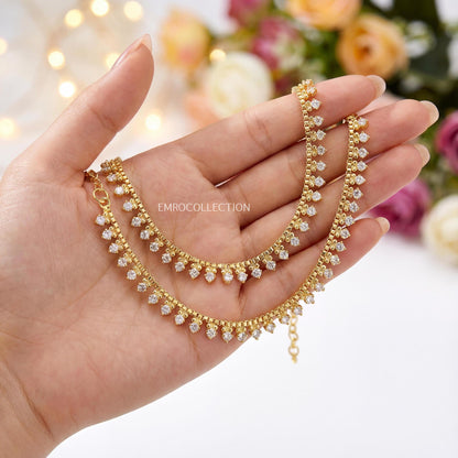 Golden anklets with diamonds held in a hand against a blurred floral background. Trending Pakistani anklets