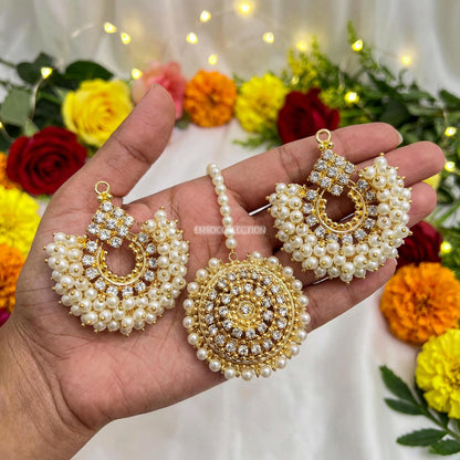 Gold and pearl earrings held in a hand with a floral background - trending pakistani chand bali earrigns tikka set