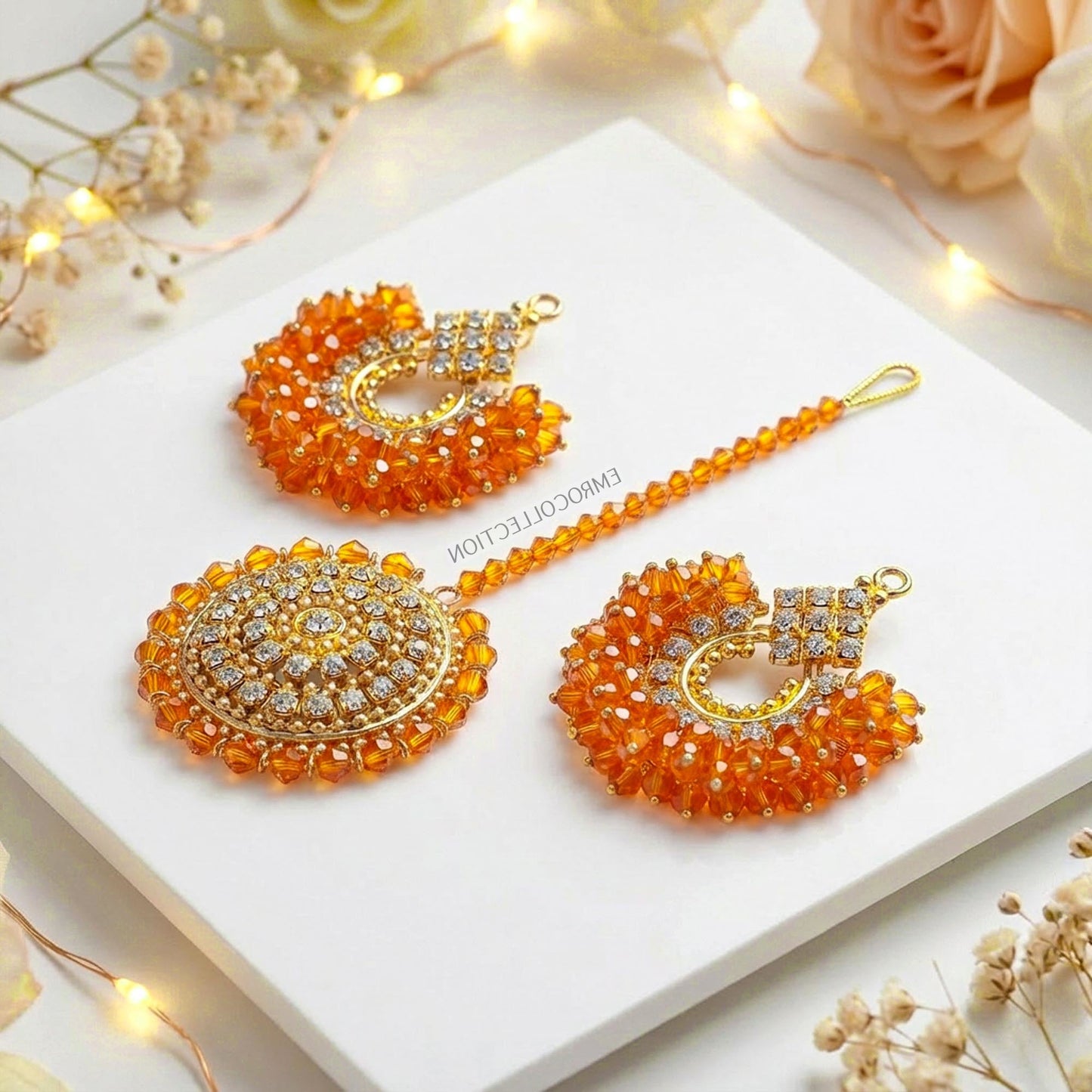 orange and gold chand bali earrings tikka set on a white surface with decorative lights and flowers.