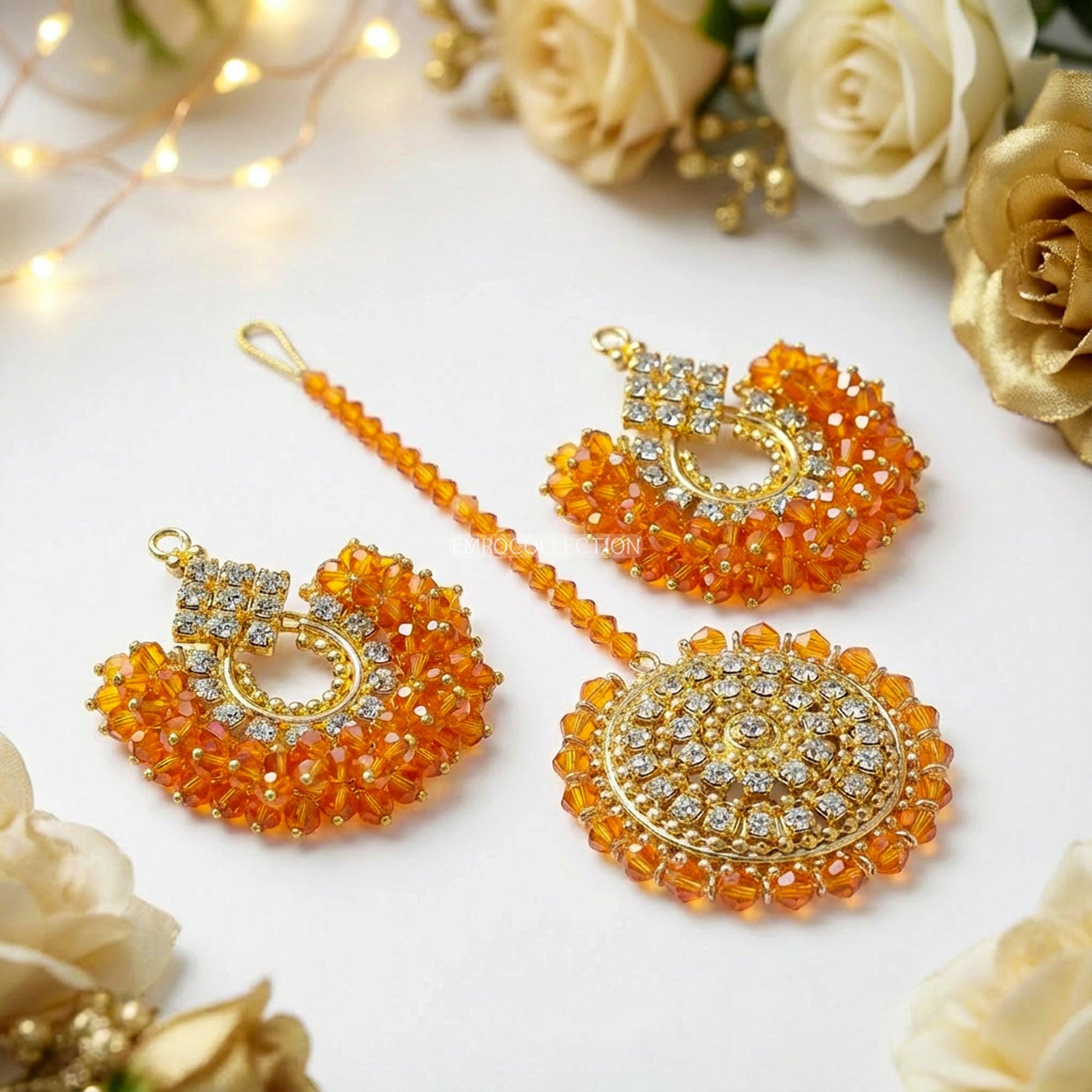 Orange and gold chand bali earrings tikka set on a white surface with decorative lights and flowers.